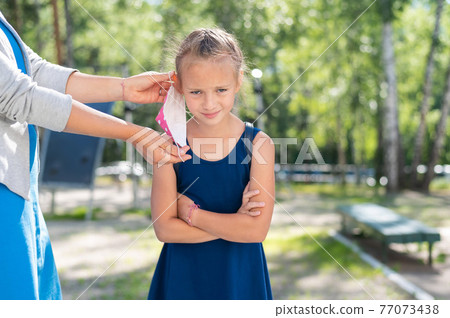Thoughtful woman wears a protective mask on a disgruntled daughter. Girl goes back to school Thoughtful woman wears a protective mask on a disgruntled daughter. Girl goes back to school 77073438
