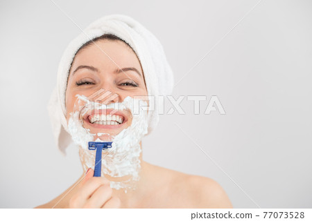 A woman with a white terry towel on her head and with shaving foam on her face shaves on a white background 77073528