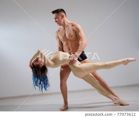 Acrobatic couple perform number on a white background. A duet of gymnasts rehearsing a performance with support. A man and a very flexible woman are dancing. Acrobatic couple perform number on a white background. A duet of gymnasts rehearsing a performance with support. A man and a very flexible woman are dancing. 77073612