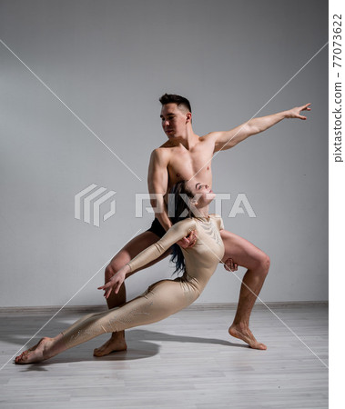 Acrobatic couple perform number on a white background. A duet of gymnasts rehearsing a performance with support. A man and a very flexible woman are dancing. Acrobatic couple perform number on a white background. A duet of gymnasts rehearsing a performance with support. A man and a very flexible woman are dancing. 77073622