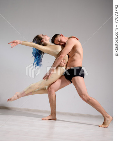 Acrobatic couple perform number on a white background. A duet of gymnasts rehearsing a performance with support. A man and a very flexible woman are dancing. Acrobatic couple perform number on a white background. A duet of gymnasts rehearsing a performance with support. A man and a very flexible woman are dancing. 77073742