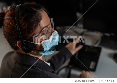 Rear view of a woman working in the office wearing a medical mask. Female call center operator wearing a protective mask. Rear view of a woman working in the office wearing a medical mask. Female call center operator wearing a protective mask. 77073743