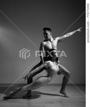A man and a woman are dancing modern ballet. Acrobatic couple perform number on a white background. A duet of gymnasts rehearsing a performance with support. Monochrome photo. A man and a woman are dancing modern ballet. Acrobatic couple perform number on a white background. A duet of gymnasts rehearsing a performance with support. Monochrome photo. 77073898
