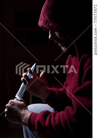 A dangerous bearded man in a red hood holds a sharp knife near his face. A criminal or a killer with knives in the dark. 77073971
