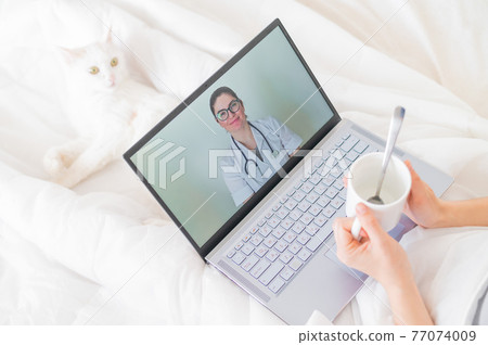 A faceless woman drinks an antipyretic and watches a video from a doctor recommendation. Online doctor consultation on a webcam without leaving your home. The cat lies in bed with a sick housewife. A faceless woman drinks an antipyretic and watches a video from a doctor recommendation. Online doctor consultation on a webcam without leaving your home. The cat lies in bed with a sick housewife. 77074009