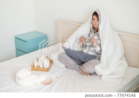 Woman in bed covered by a blanket with a thermometer in her hands. A girl with a fever is sitting in the bedroom with a laptop on her knees. White cat next to the owner Woman in bed covered by a blanket with a thermometer in her hands. A girl with a fever is sitting in the bedroom with a laptop on her knees. White cat next to the owner 77074148