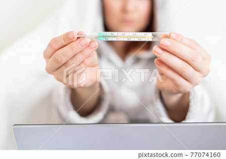 A woman in pajamas with a fever shows a mercury thermometer at the camera. Faceless blurry girl with fever is sitting on the bed. A woman in pajamas with a fever shows a mercury thermometer at the camera. Faceless blurry girl with fever is sitting on the bed. 77074160