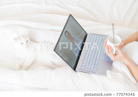Unrecognizable girl watching a video with medical advice on a laptop. A woman conducts an online conversation with a doctor on a webcam. The cat lies in the legs of a cold owner. 77074348