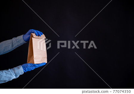 Female hands in a blue glove holds a brown paper bag on a black background. Safe food delivery to your home. A courier in a denim shirt holds out a craft cardboard bag to a customer. Female hands in a blue glove holds a brown paper bag on a black background. Safe food delivery to your home. A courier in a denim shirt holds out a craft cardboard bag to a customer. 77074350