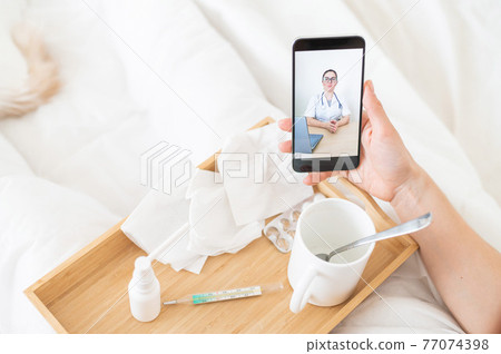 Telemedicine concept. Close-up of a doctor on a smartphone display during an online medical consultation with a patient. Wooden tray with medicines and a thermometer on the patient's bed. Telemedicine concept. Close-up of a doctor on a smartphone display during an online medical consultation with a patient. Wooden tray with medicines and a thermometer on the patient's bed. 77074398