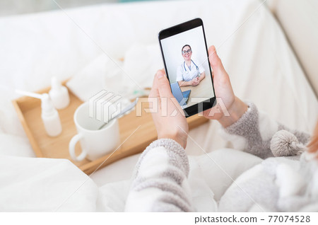 Woman in bed in pajamas with a smartphone in her hands. A flu patient is watching a medical video blog. A friendly doctor gives an online consultation by phone. 77074528