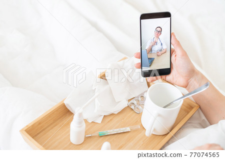 Female hands holding a phone. Remote appointment with a doctor. A woman is talking to the attending physician from home. Medications and thermometer on a wooden tray. Female hands holding a phone. Remote appointment with a doctor. A woman is talking to the attending physician from home. Medications and thermometer on a wooden tray. 77074565