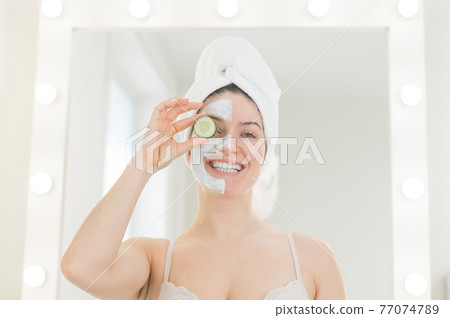 Cheerful woman with a towel on her hair and in a clay face mask fooling around with cucumbers in her hands. Taking care of beauty at home Cheerful woman with a towel on her hair and in a clay face mask fooling around with cucumbers in her hands. Taking care of beauty at home 77074789
