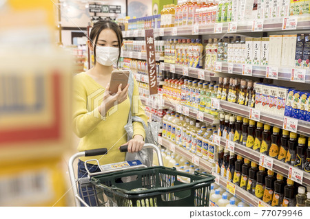 在一家超市購物場景中看著智能手機的面具的女性購物者 77079946