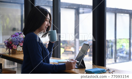 Side view of young woman working on her project with computer tablet and drinking coffee while sitting at workstation. 77093154