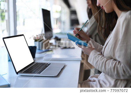 Cropped shot of young female sitting in front of her laptop and making some note in office. Cropped shot of young female sitting in front of her laptop and making some note in office. 77097072