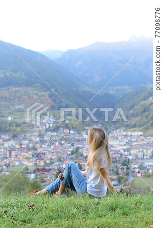 Young woman sitting in mountains, little village in background. Young woman sitting in mountains, little village in background. 77098776