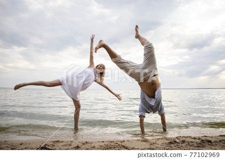 Creative couple relaxing on the ocean beach. A man and a woman do somersaults and jumps in the water. The concept of unusual couples and ingenuity in poses for a photo Creative couple relaxing on the ocean beach. A man and a woman do somersaults and jumps in the water. The concept of unusual couples and ingenuity in poses for a photo 77102969