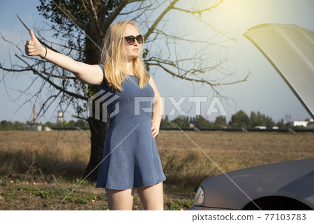 A blonde in a blue sundress votes on the road, near her car. An open bonnet indicates a breakdown in the vehicle. The concept of helping motorists in a difficult situation. Sweet girl in trouble 77103783