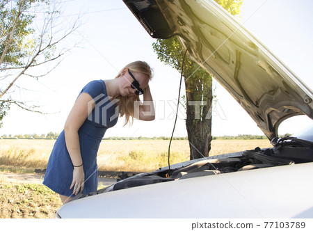The blonde girl looks at the engine, which does not work. The woman touches the parts under the hood, but does not understand the cause of the breakdown. Concept of unpleasant situations on the road 77103789