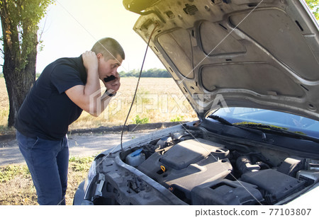 An angry man looks at the idle car engine. The driver tries to explain the cause of the breakdown to the mechanic using his smartphone and call for help. Concept of unpleasant situations on the road An angry man looks at the idle car engine. The driver tries to explain the cause of the breakdown to the mechanic using his smartphone and call for help. Concept of unpleasant situations on the road 77103807