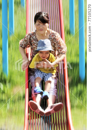 A 3-year-old boy playing with his grandma on a park slide 77105270