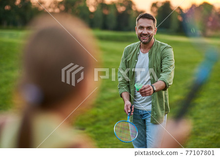 Happy father holding badminton racket and shuttlecock while playing with his little daughter outdoors in the park on a summer day Happy father holding badminton racket and shuttlecock while playing with his little daughter outdoors in the park on a summer day 77107801