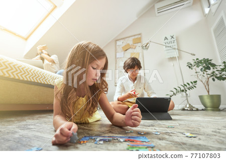 Family at home. Little girl lying on the floor in the living room and playing with puzzles while her brother using digital tablet and doing homework 77107803