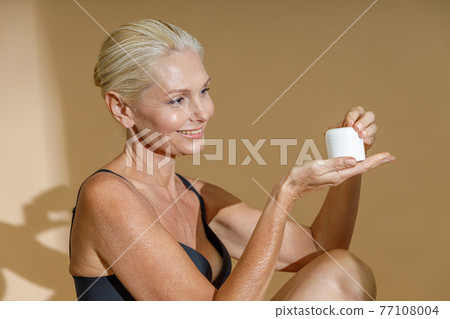 Smiling mature woman in black bra smiling and looking at white jar of cream beauty product in her hands, posing half naked isolated over beige background Smiling mature woman in black bra smiling and looking at white jar of cream beauty product in her hands, posing half naked isolated over beige background 77108004