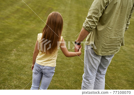 Back view of a little girl holding hand of her father while spending time together outdoors Back view of a little girl holding hand of her father while spending time together outdoors 77108070