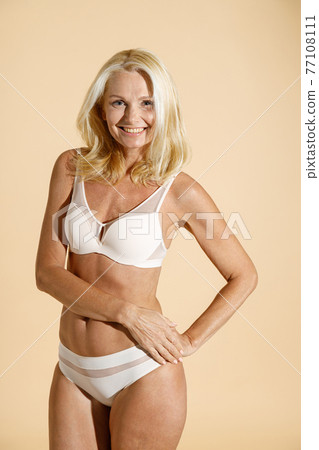 Cheerful caucasian mature blonde woman in lingerie smiling at camera, standing isolated over beige background, posing half naked Cheerful caucasian mature blonde woman in lingerie smiling at camera, standing isolated over beige background, posing half naked 77108111