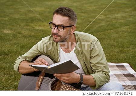 Young thoughtful man wearing eyeglasses holding acoustic guitar and composing a song while sitting on a green grass outdoors 77108171