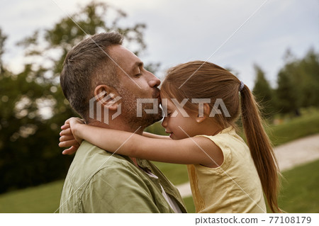 Young loving father kissing his cute little daughter in forehead while spending time together outdoors on a summer day, sitting on a green grass in park Young loving father kissing his cute little daughter in forehead while spending time together outdoors on a summer day, sitting on a green grass in park 77108179