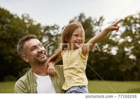 Cute and happy little girl spending time with her young loving father while visiting park on a warm summer day, she is pointing at something and smiling Cute and happy little girl spending time with her young loving father while visiting park on a warm summer day, she is pointing at something and smiling 77108208