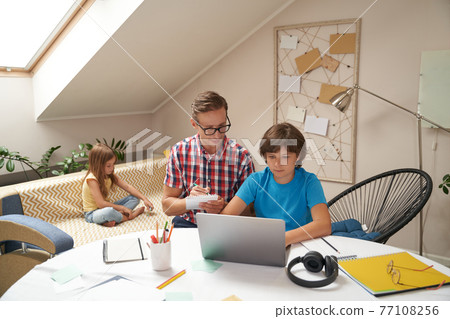 Dad helping his son with school homework, boy learning online while his little sister sitting on sofa on the background 77108256