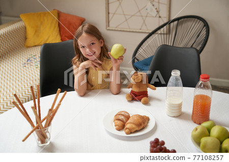 Happy caucasian girl sitting at the table at home, holding green apple and smiling at camera, having breakfast at home 77108257
