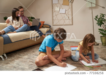 Little boy and girl sitting on the wooden floor and drawing with colorful pencils while their parents, happy mother and father relaxing on sofa and using laptop Little boy and girl sitting on the wooden floor and drawing with colorful pencils while their parents, happy mother and father relaxing on sofa and using laptop 77108258