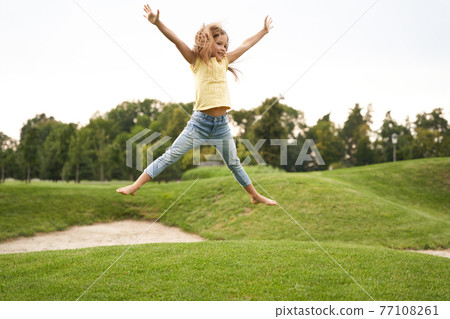 Happy childhood concept. Cute little girl in casual clothes jumping and having fun while visiting park on a sunny summer day, she is smiling and feeling so excited Happy childhood concept. Cute little girl in casual clothes jumping and having fun while visiting park on a sunny summer day, she is smiling and feeling so excited 77108261