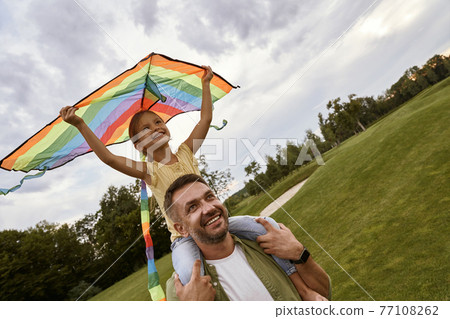 Excited little girl sitting on shoulders of her father and playing with colorful kite, daughter and daddy spending time together in park on a warm day 77108262