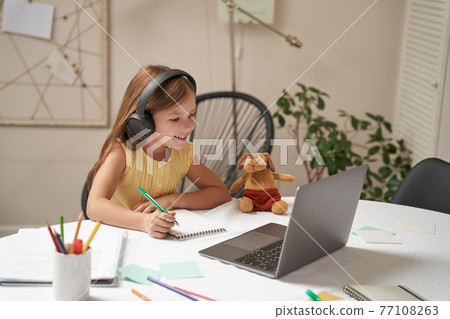 E-learning system. Smart little caucasian girl in headphones sitting at the table, using laptop and watching video lesson 77108263