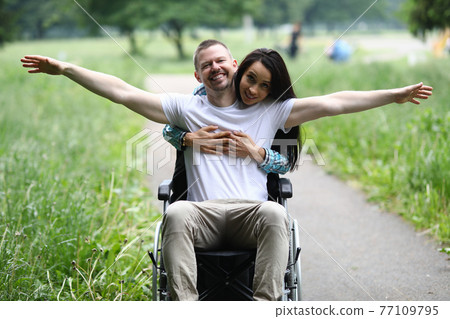 Woman hugs joyful man in wheelchair in park Woman hugs joyful man in wheelchair in park 77109795