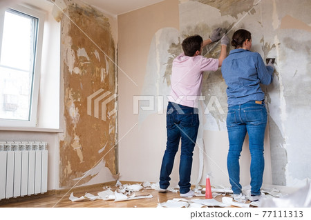 Young couple removing old wallpaper from walls preparing for flat renovation. 77111313