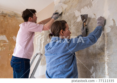 Young couple removing old wallpaper from walls preparing for flat renovation. 77111335