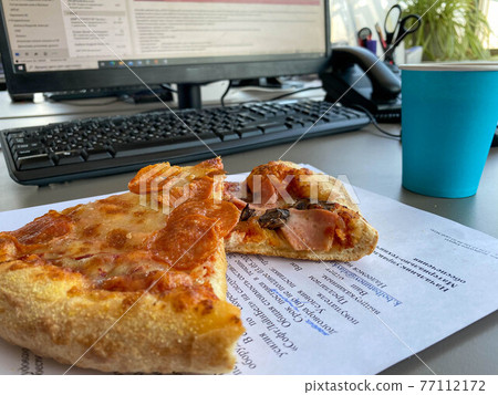 Delicious nourishing beautiful appetizing pizza with piperone sausage lies on a working business desktop in front of a computer in the office. A quick lunch for an office worker 77112172