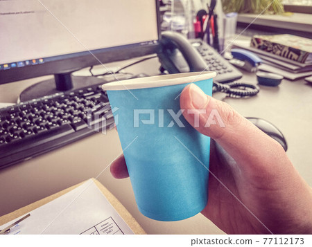 Cardboard blue coffee cup from a coffee shop on a desktop with a computer with a keyboard and mouse and office supplies of an office worker during lunch. Business coffee break 77112173