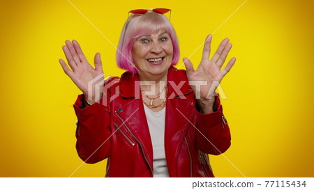 Happy friendly elderly woman waves hand palm in hello gesture welcomes someone positive emotions 77115434