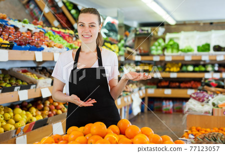 Smiling woman seller wearing apron standing near fresh oranges 77123057