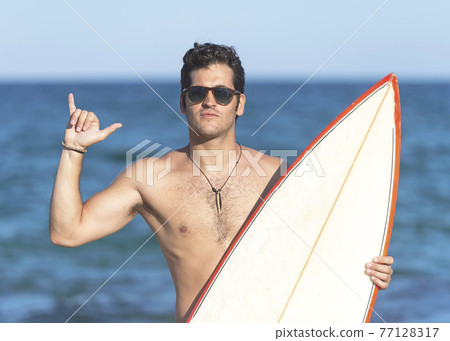 Surfer guy happy with surf surfing smiling doing hawaiian shaka hand sign for fun during surf session in ocean waves on beach vacation. Surfer guy happy with surf surfing smiling doing hawaiian shaka hand sign for fun during surf session in ocean waves on beach vacation. 77128317