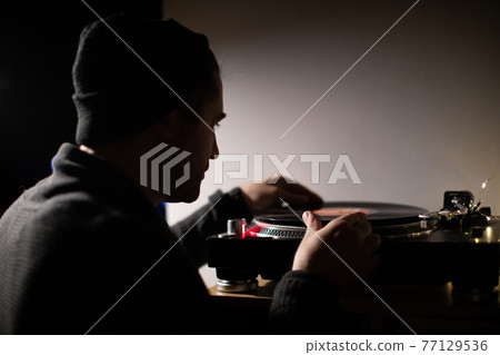 Young unshaven handsome millennial guy puts record on turntable backlit 77129536