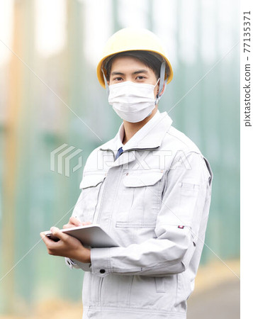 Men in work clothes wearing a mask and holding a tablet Men in work clothes wearing a mask and holding a tablet 77135371
